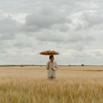 A Woman in a Beige Coat Holding an Umbrella while Standing in a Rye Field