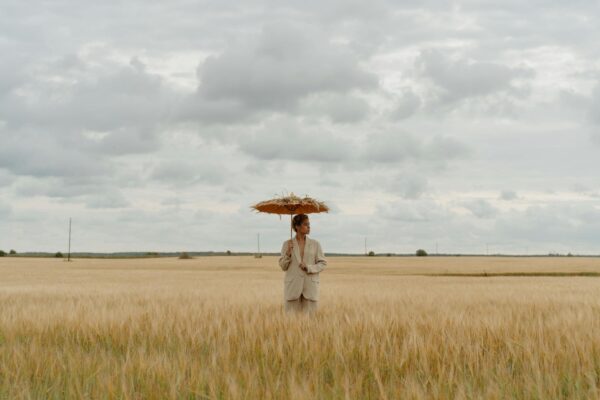 A Woman in a Beige Coat Holding an Umbrella while Standing in a Rye Field